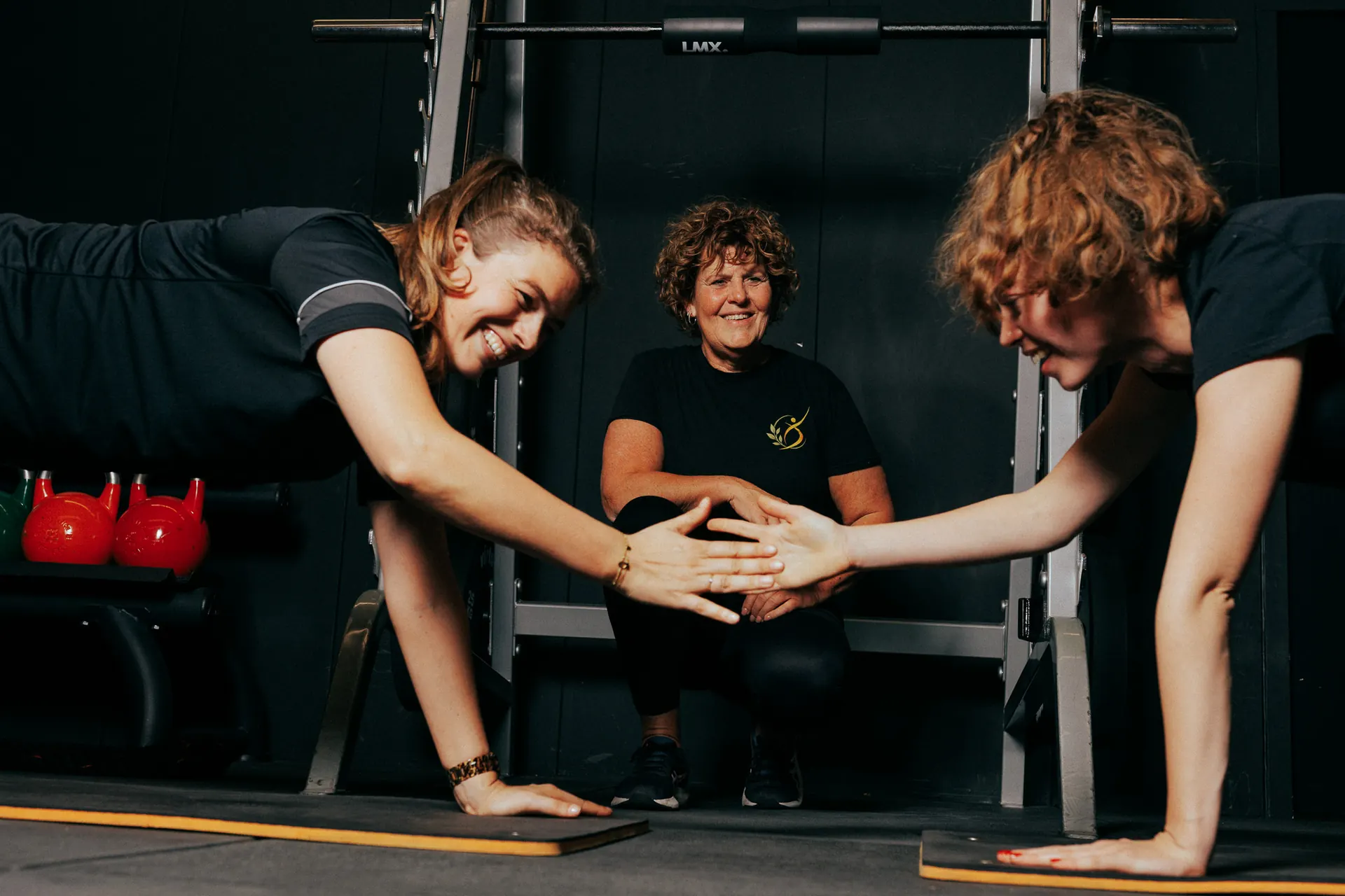 Twee sporters geven elkaar een high five in plankpositie, met Carla op de achtergrond, bij LevelFit Enschede